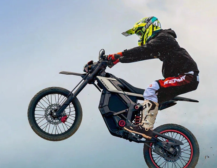 Wearing protective gear, a rider launches mid-air on the Segway Xaber 300 electric dirt bike by Segway, highlighting its off-road capabilities against a backdrop of clouds.
