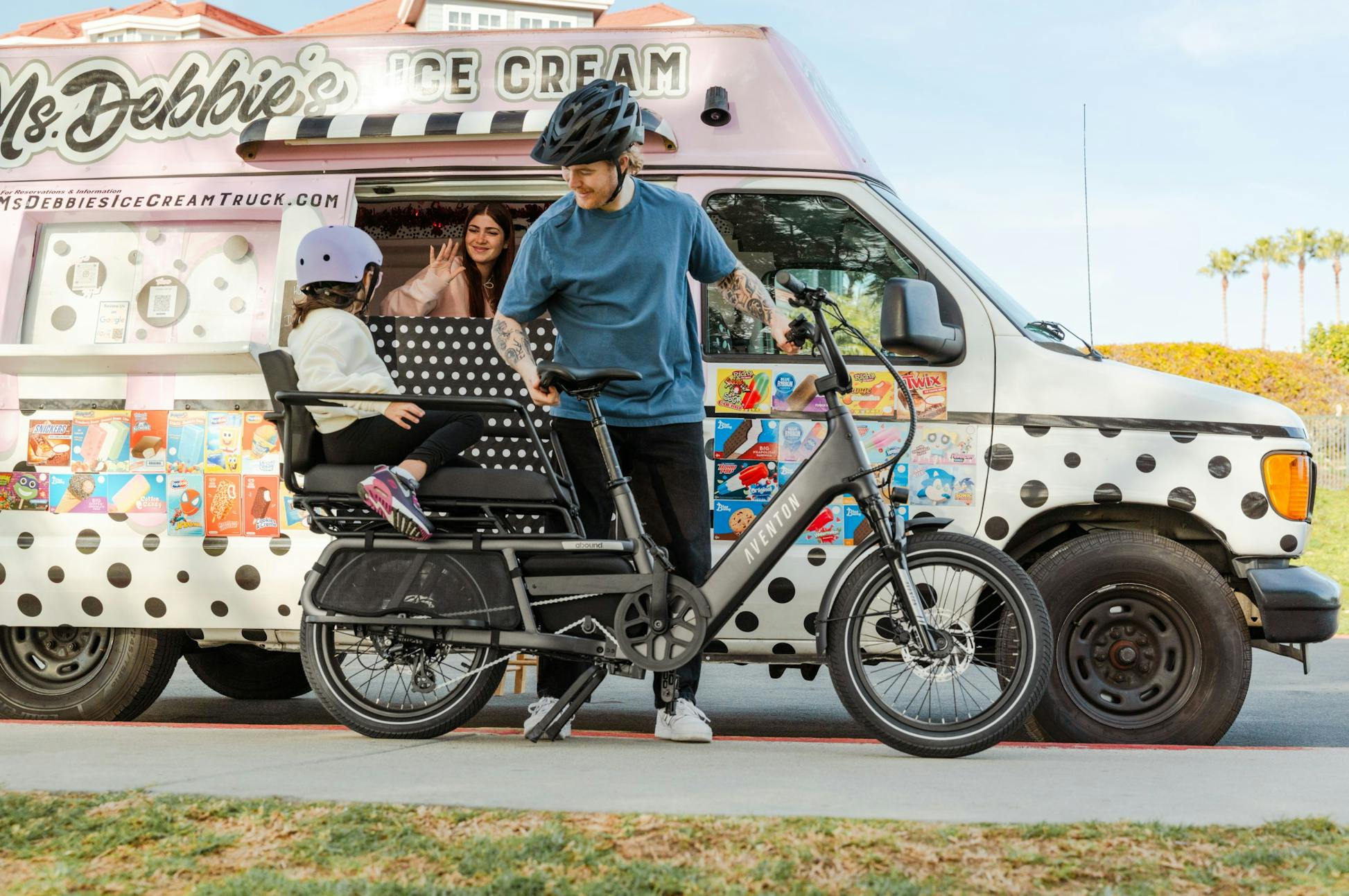 A man and child ride up to an ice cream truck on the Aventon Abound LR by Aventon, where a woman inside serves them a treat through the window.