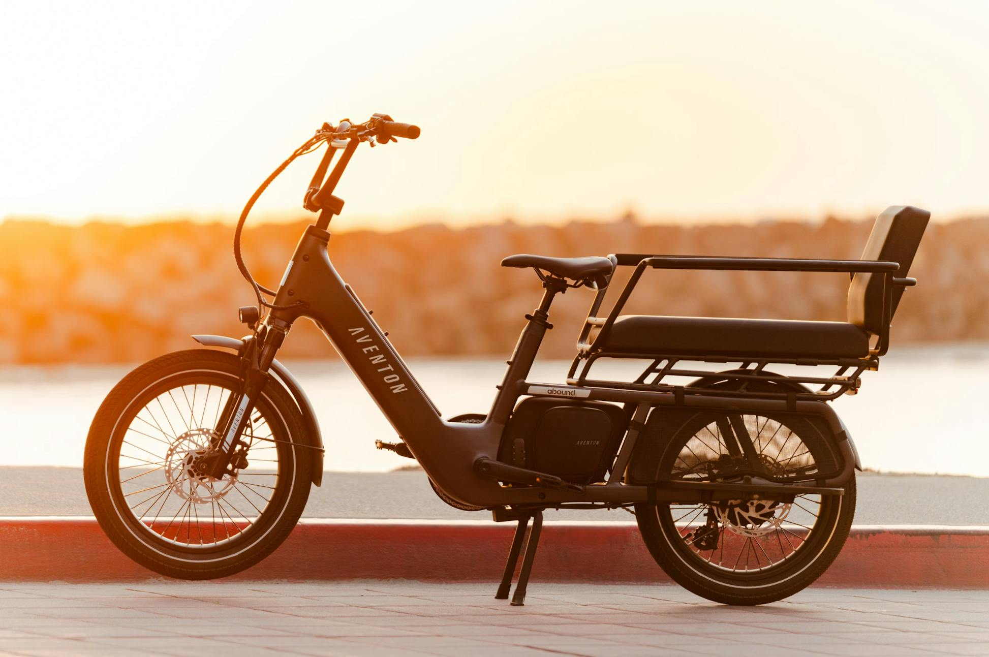 A black Aventon Abound LR electric cargo bike from Aventon with a rear bench seat is parked on a sidewalk near the water at sunset.