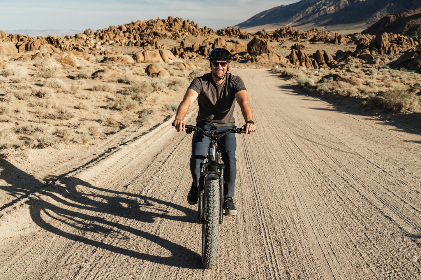 A person wearing a helmet rides a Tampa Bay eBikes Aventon - Aventure M fat tire eBike on a dirt road through a rocky desert landscape under a clear sky.