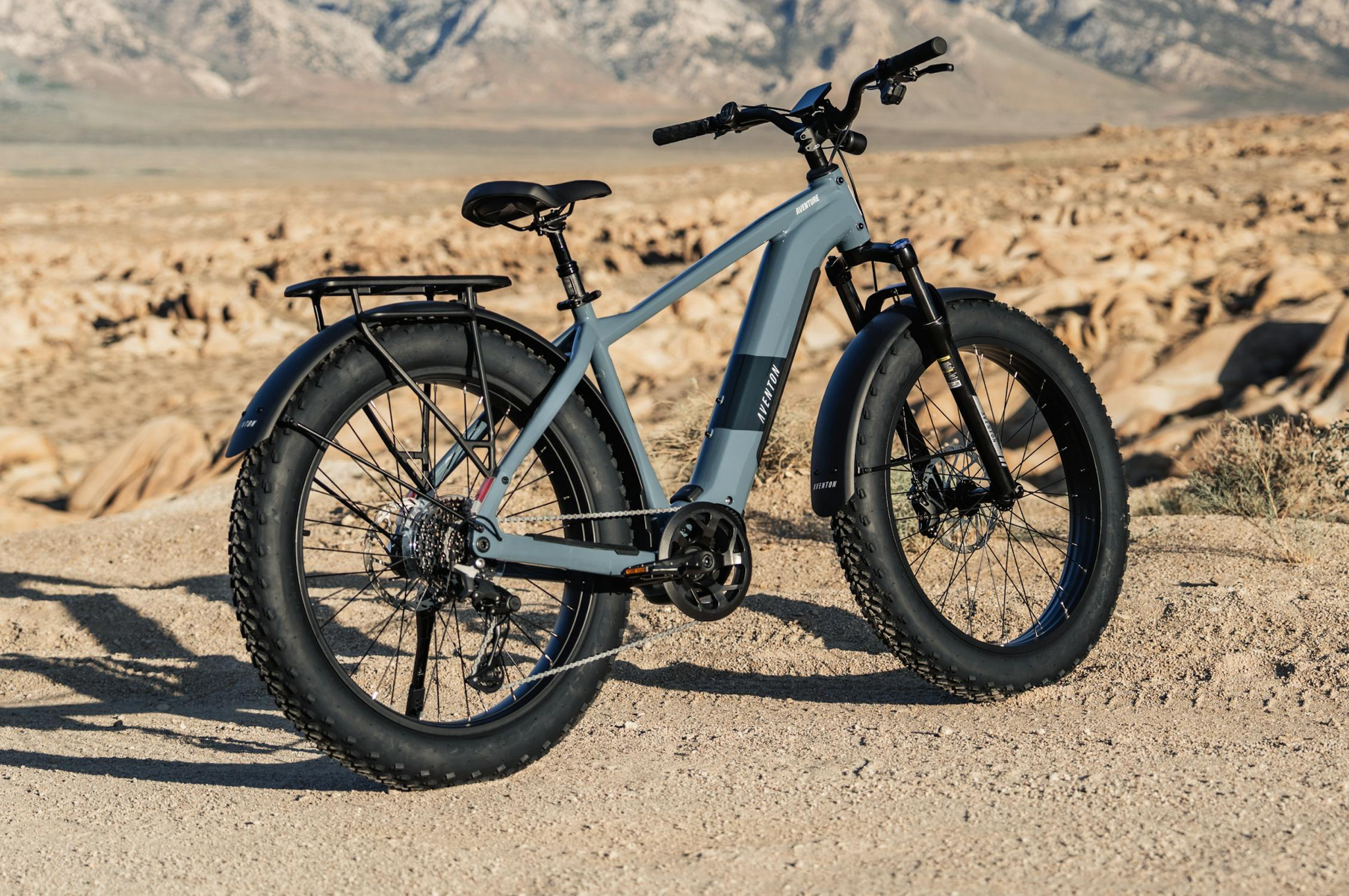 A gray Aventon - Aventure M fat-tire electric bike from Tampa Bay eBikes is parked on a dirt path in a rocky, mountainous desert landscape.
