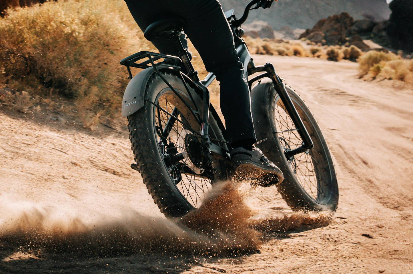 A rider carves a dusty path on the Aventon - Aventure M from Tampa Bay eBikes, sand flying as they turn past desert plants and rocky hills—a high-torque fat tire eBike built for adventurous off-road rides.
