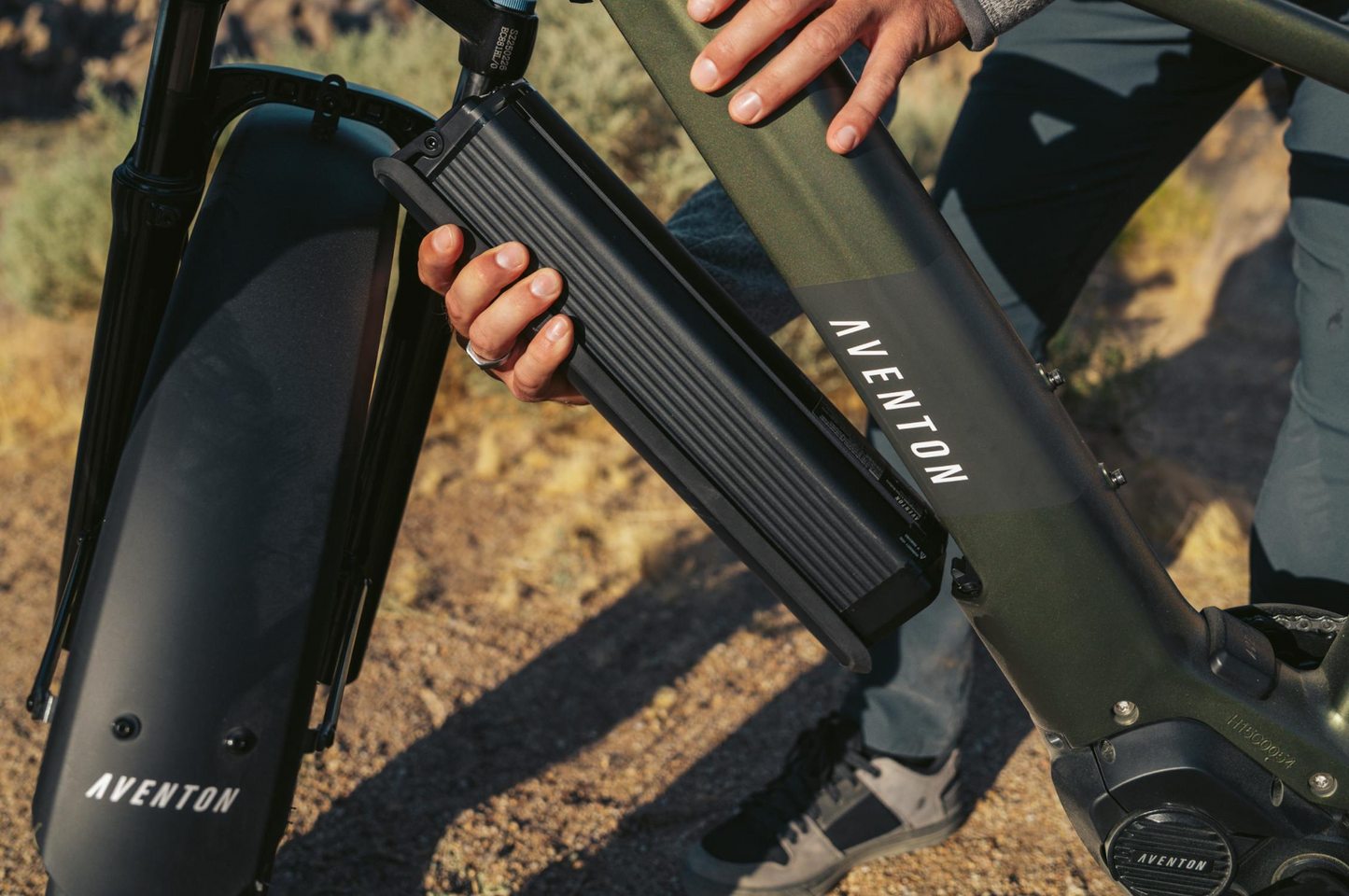 A person inserts a rectangular battery into the frame of the Tampa Bay eBikes Aventon - Aventure M, preparing to enjoy high-torque rides on any terrain outdoors.