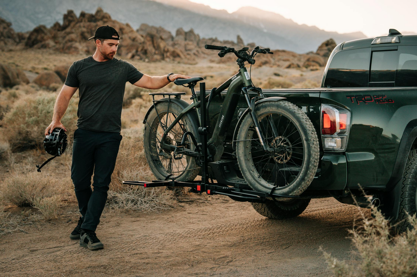 A man stands beside a pickup truck with mountain bikes, including a Tampa Bay eBikes Aventon Aventure M fat tire eBike, mounted on a rear rack in a dry, rocky landscape.
