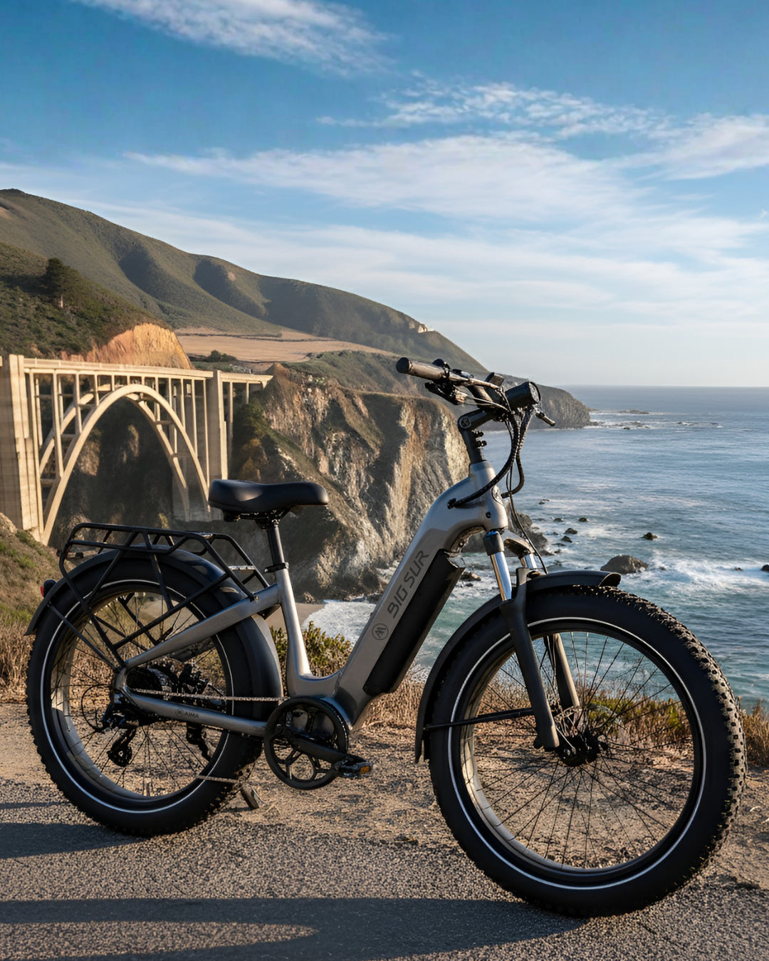 The AIMA - Big Sur 2.0 electric bike from AIMA is parked on a coastal road with ocean views, a large concrete arch bridge, and dramatic cliffs in the background.