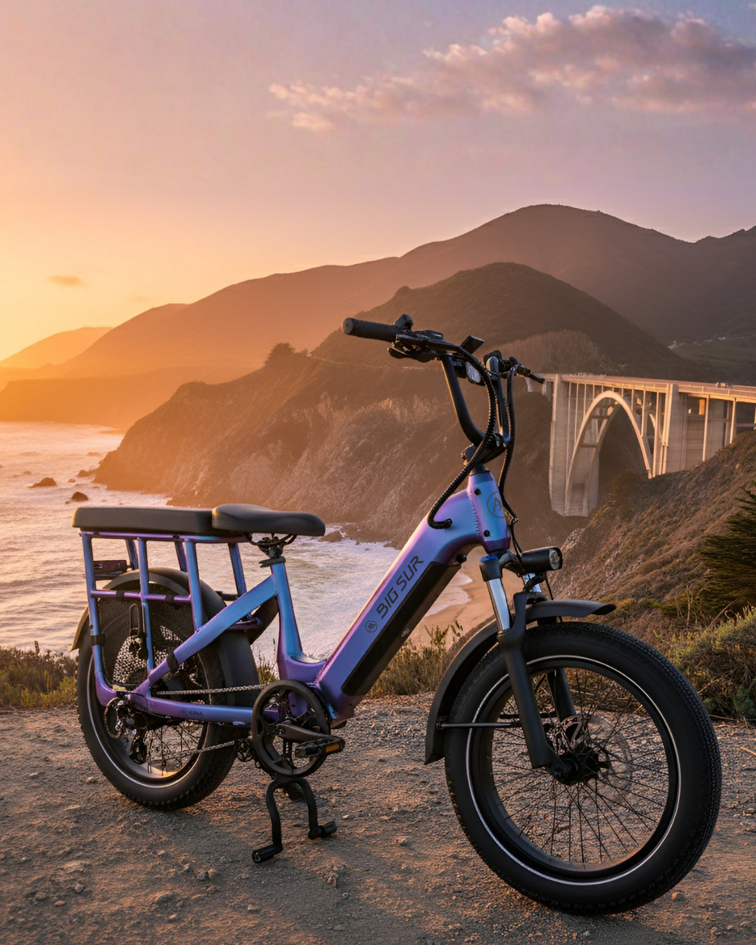 An AIMA Big Sur U Cargo eBike in purple is parked on a dirt path overlooking the ocean, mountains, and a coastal bridge at sunset.