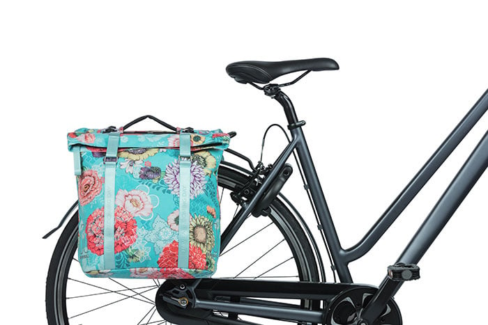 The Basil Bloom Field Double Pannier by Basil, featuring a turquoise floral pattern and made from recycled PET, is mounted on the rear rack of a black bicycle against a white background.