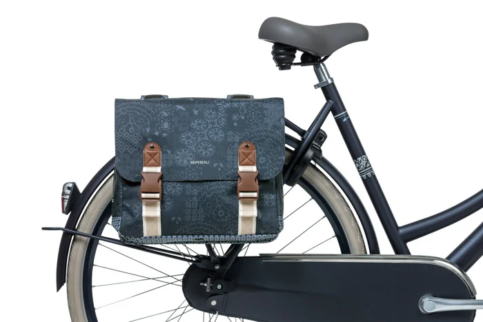 A black bicycle with a Basil Boheme Double Pannier Bag by Basil in a gray pattern, attached to the rear rack and featuring brown straps and silver buckles.