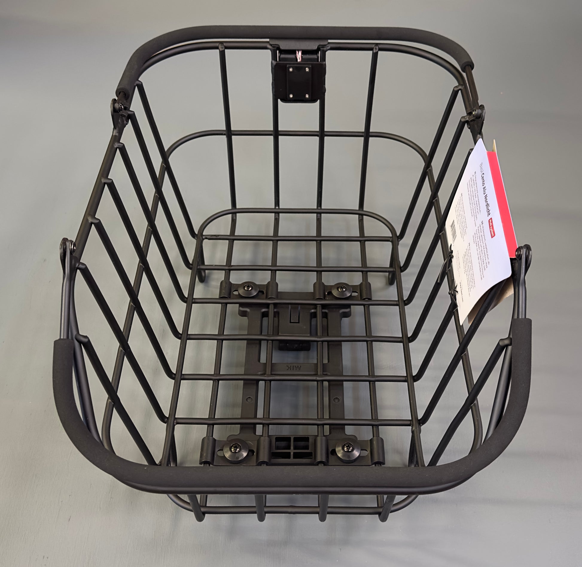 The Basil Cento MIK Rear Basket is a black metal wire e-bike basket with a mounting bracket at the base and a paper tag attached. Empty and placed on a plain surface, it is compatible with the MIK system for easy e-bike installation.