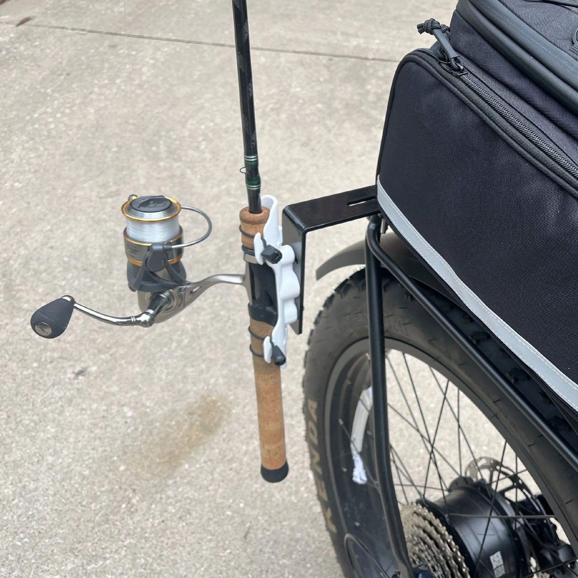 A Bikase Fishing Rod Holder secures a fishing rod to the rear rack of a bicycle parked on concrete.