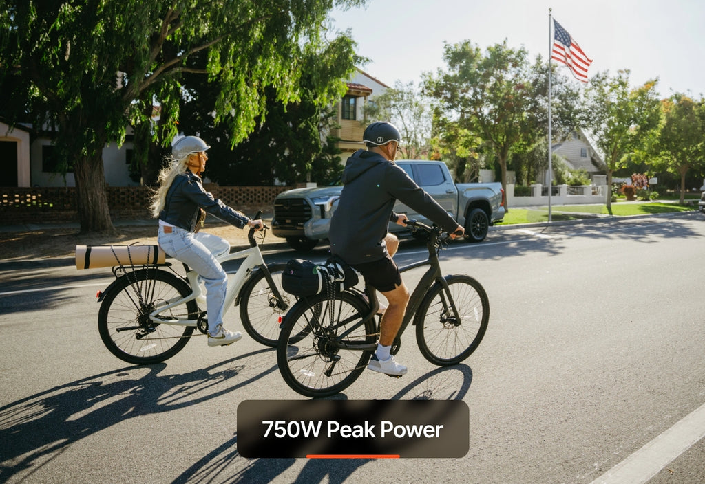 Two people ride Urtopia Carbon Classic bikes on a suburban street in daylight, both wearing helmets. An American flag waves in the background. The text reads “750W Peak Power.”.