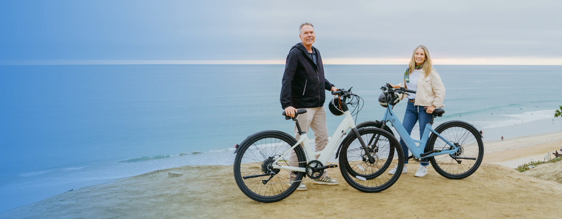 Two people with their Urtopia Carbon Classic Step-Thru eBikes stand on a cliff overlooking the ocean, with a sandy beach and calm water in the background.