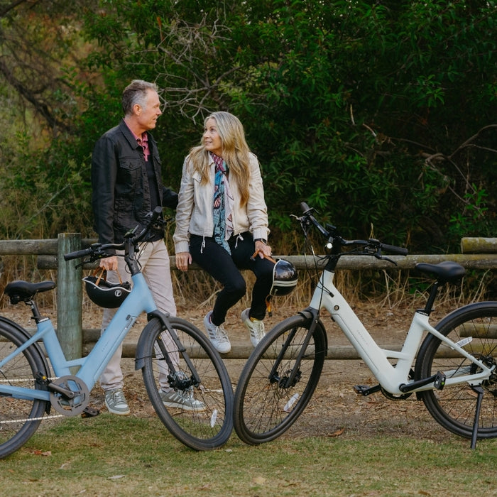 A man and a woman with bicycles stand and sit by a wooden fence in a park, smiling at each other, their Urtopia Carbon Classic Step-Thru eBikes from Urtopia resting nearby.