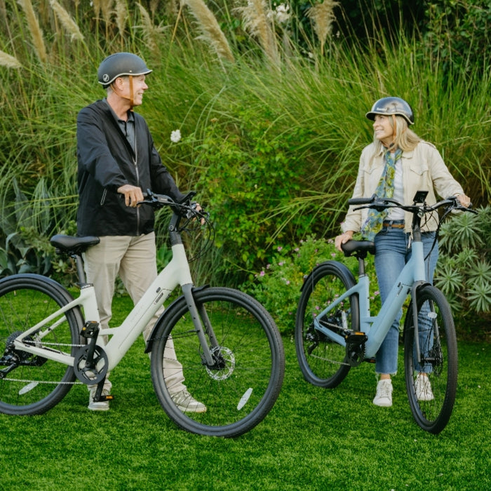 A man and woman wearing helmets stand on grass, smiling at each other with their Urtopia Carbon Classic Step-Thru eBikes by Urtopia; tall grass and greenery form the background.