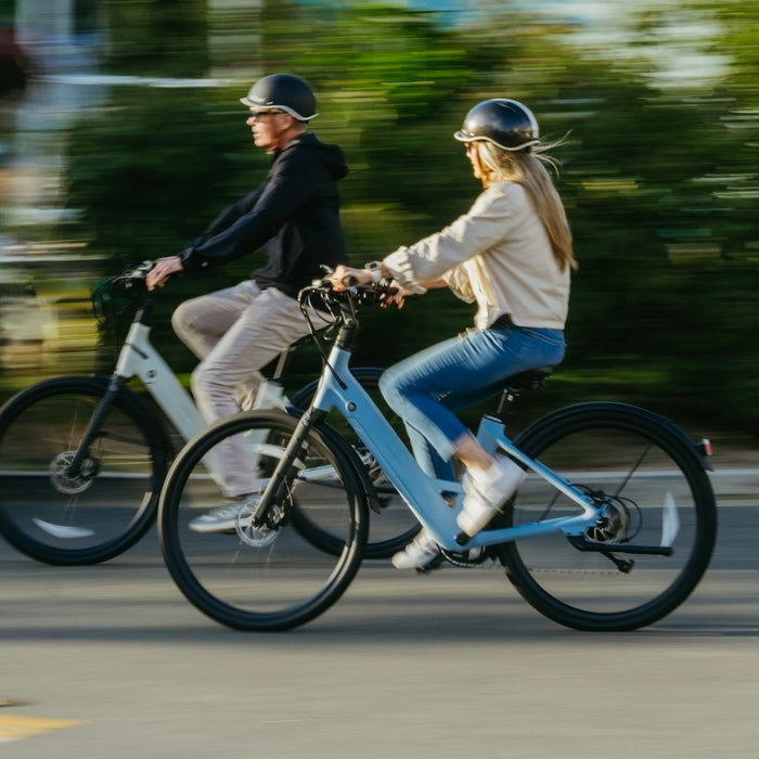 Two people wearing helmets ride Urtopia Carbon Classic Step-Thru eBikes by Urtopia on a city street, with motion blur in the background conveying their speed.