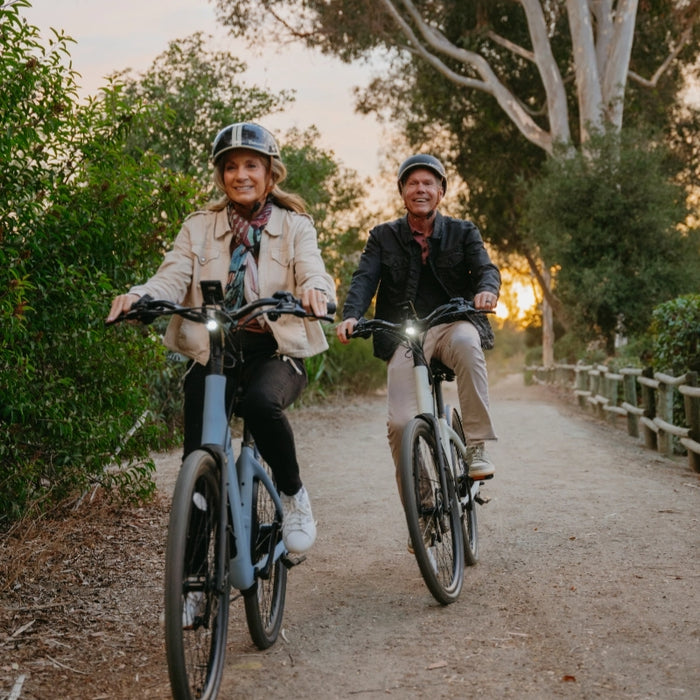 Two adults, both wearing helmets, ride Urtopia Carbon Classic Step-Thru eBikes along a dirt path lined with trees as the sun sets in the background.