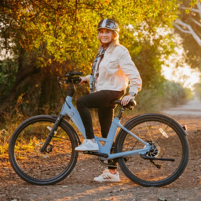 A person in a helmet and casual clothes sits on a blue Urtopia Carbon Classic Step-Thru eBike with a removable Samsung battery, riding on a dirt path among trees at sunset.