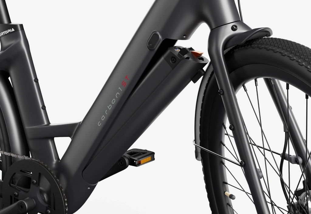 Close-up of the Urtopia Carbon Classic Step-Thru eBike's lightweight carbon frame with a removable Samsung battery, pedal, tire, and drivetrain visible, set against a white background. Brand: Urtopia.