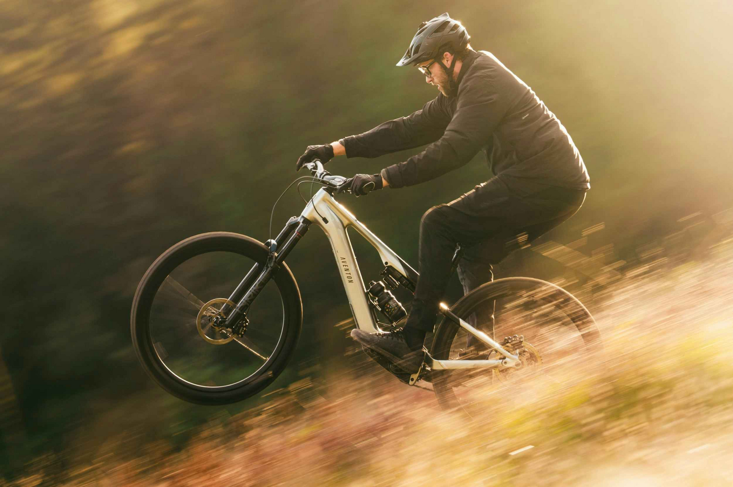 Wearing a helmet and dark clothes, a rider performs a wheelie on an Aventon Current EXP electric mountain bike by Aventon along an outdoor trail.