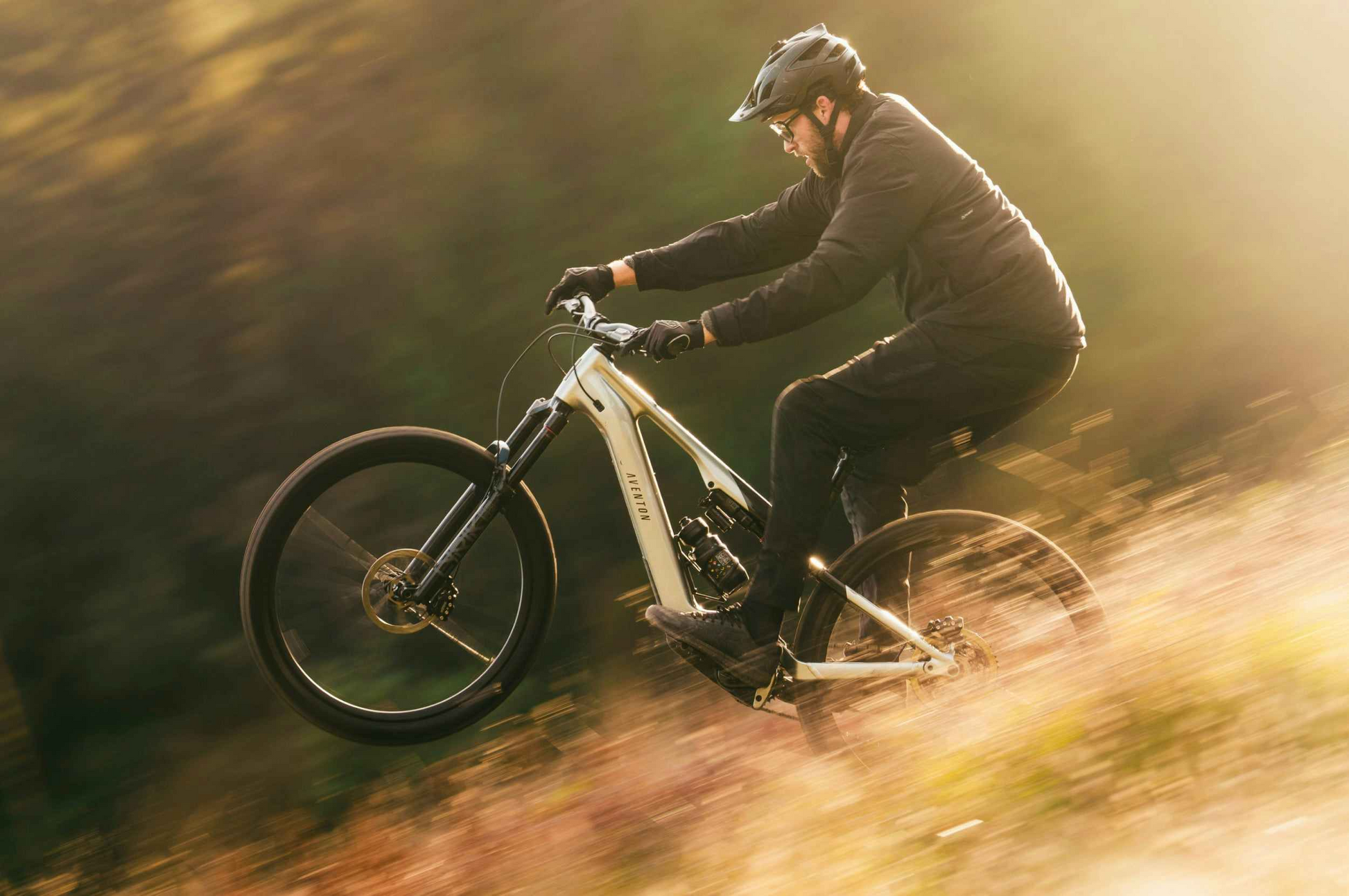 Wearing a helmet and dark clothes, a rider performs a wheelie on an Aventon Current EXP electric mountain bike by Aventon along an outdoor trail.