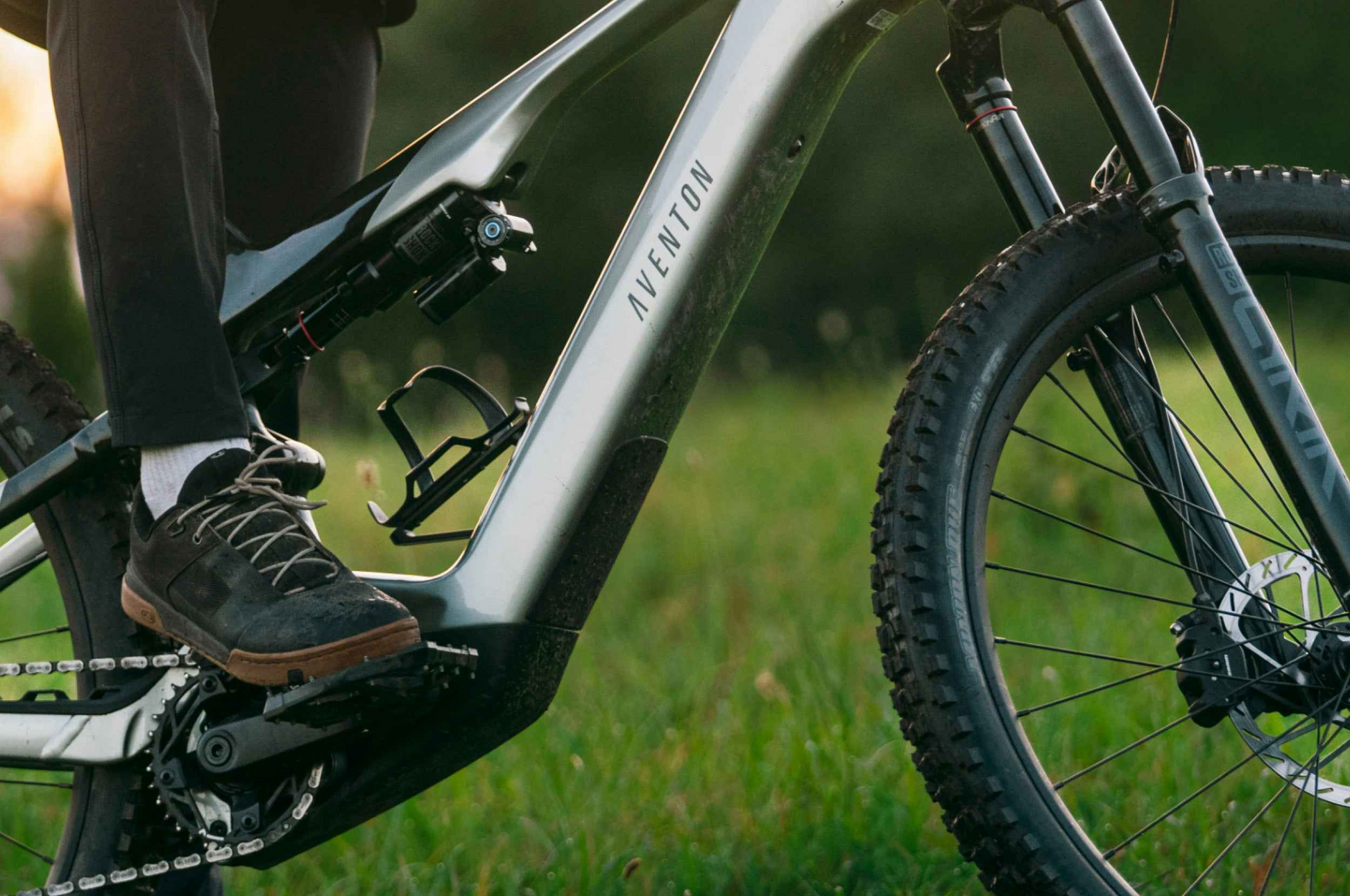 Close-up of a person’s foot on the pedal of an Aventon Current EXP electric mountain bike by Aventon, with grass and greenery in the background.