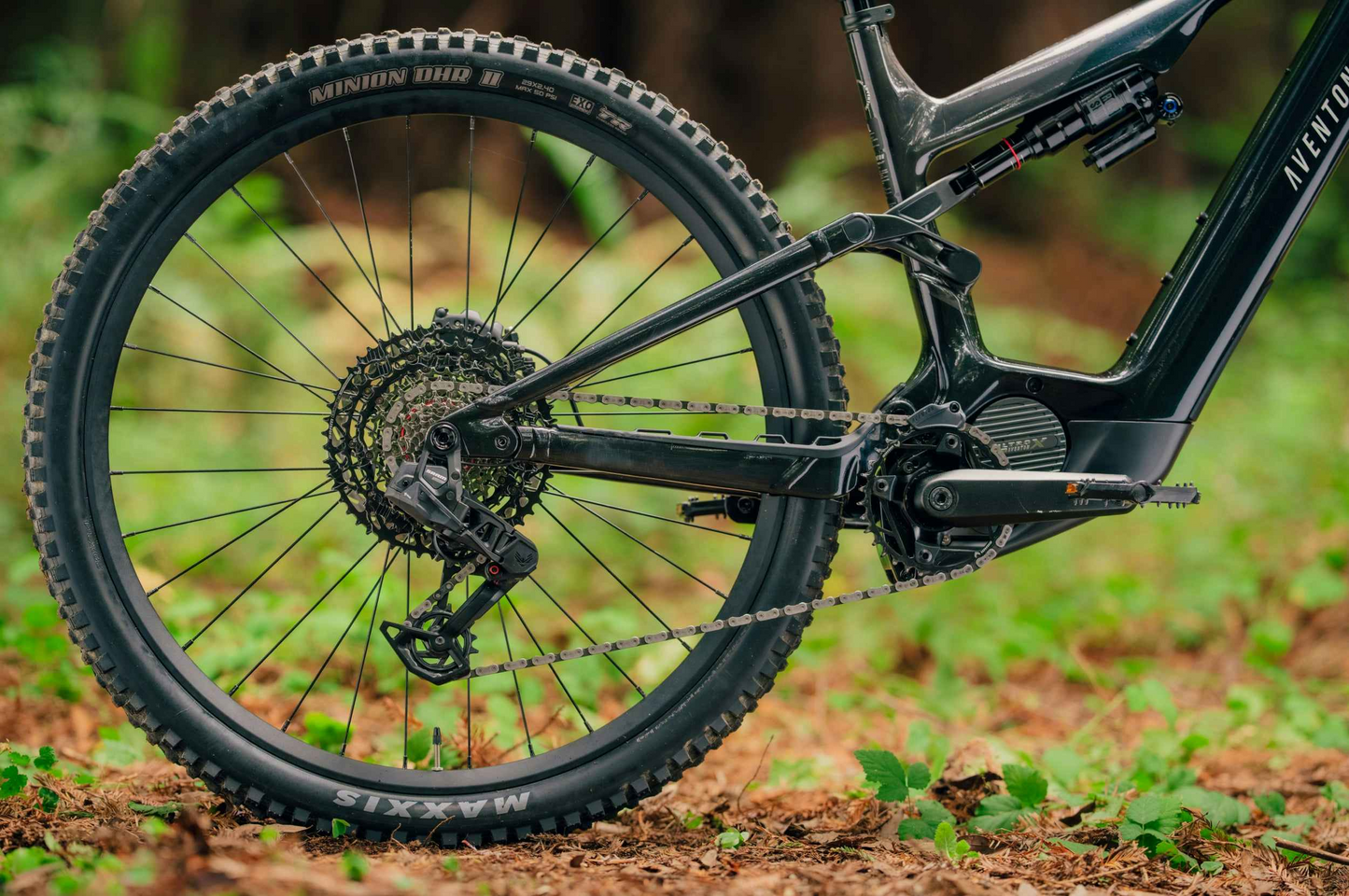 Close-up of the drivetrain and rear wheel of the Aventon Current EXP, a black carbon eMTB by Aventon with Maxxis tires, photographed outdoors on dirt and grass.