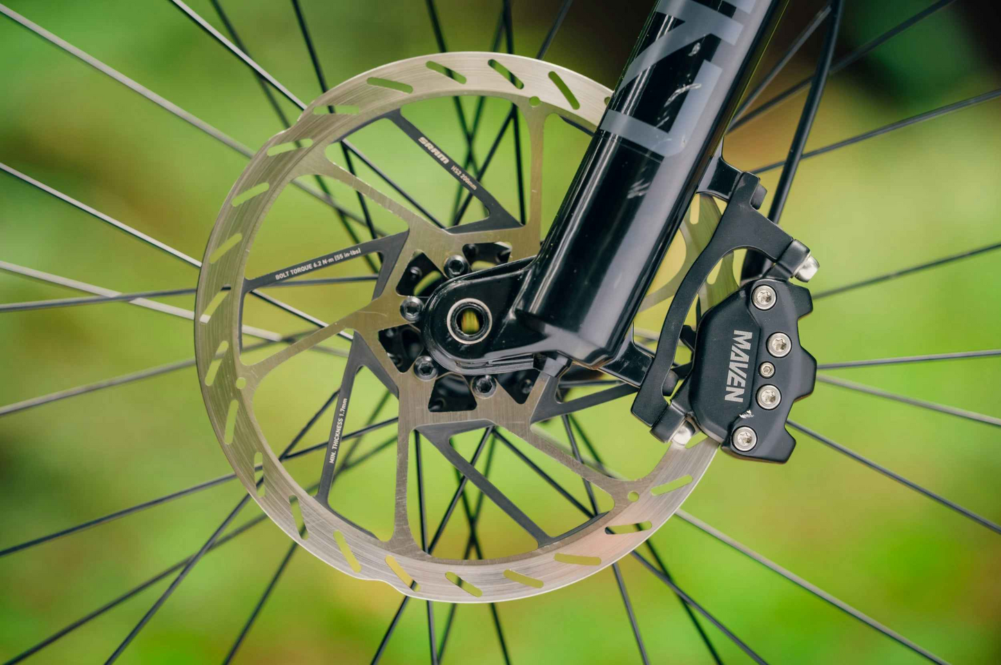 Close-up of the front disc brake system on the Aventon Current EXP carbon eMTB by Aventon, highlighting the rotor attached to the wheel hub and a "MAVEN" caliper mounted on the fork.