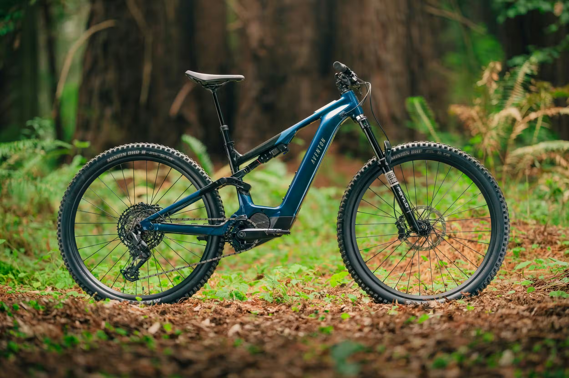 The Aventon Current ADV, a blue full-suspension eMTB by Aventon, stands on a forest floor with tall trees and lush green foliage in the background.