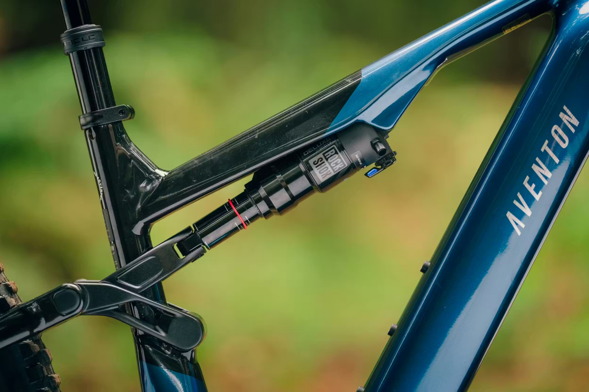 Close-up of the Aventon Current ADV by Aventon, a full-suspension eMTB, focusing on its rear suspension and shock absorber with blurred greenery in the background.