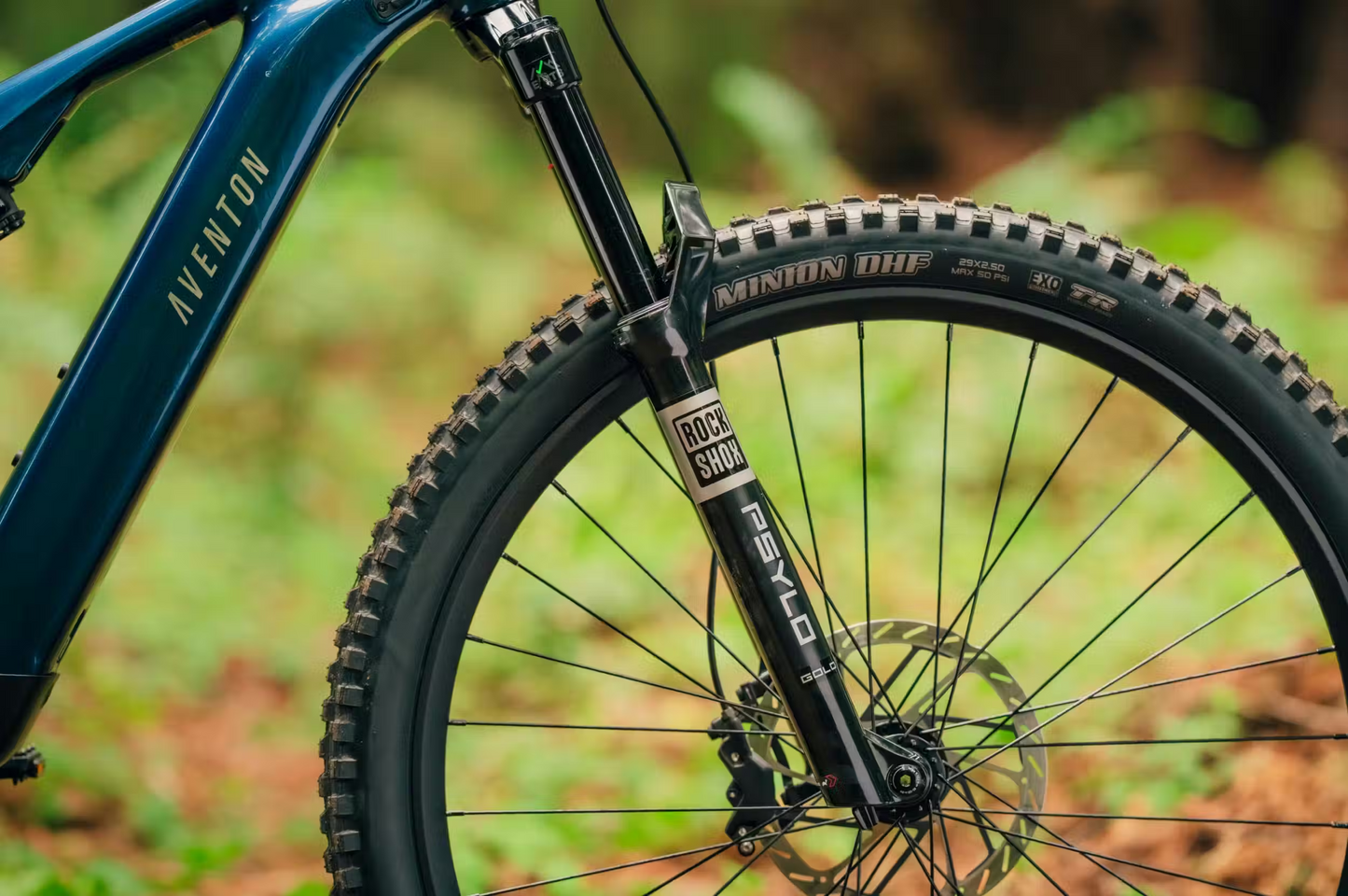 Close-up of the Aventon Current ADV's front wheel, showcasing a RockShox suspension fork and Maxxis Minion DHR tire, set against a blurred forest background.