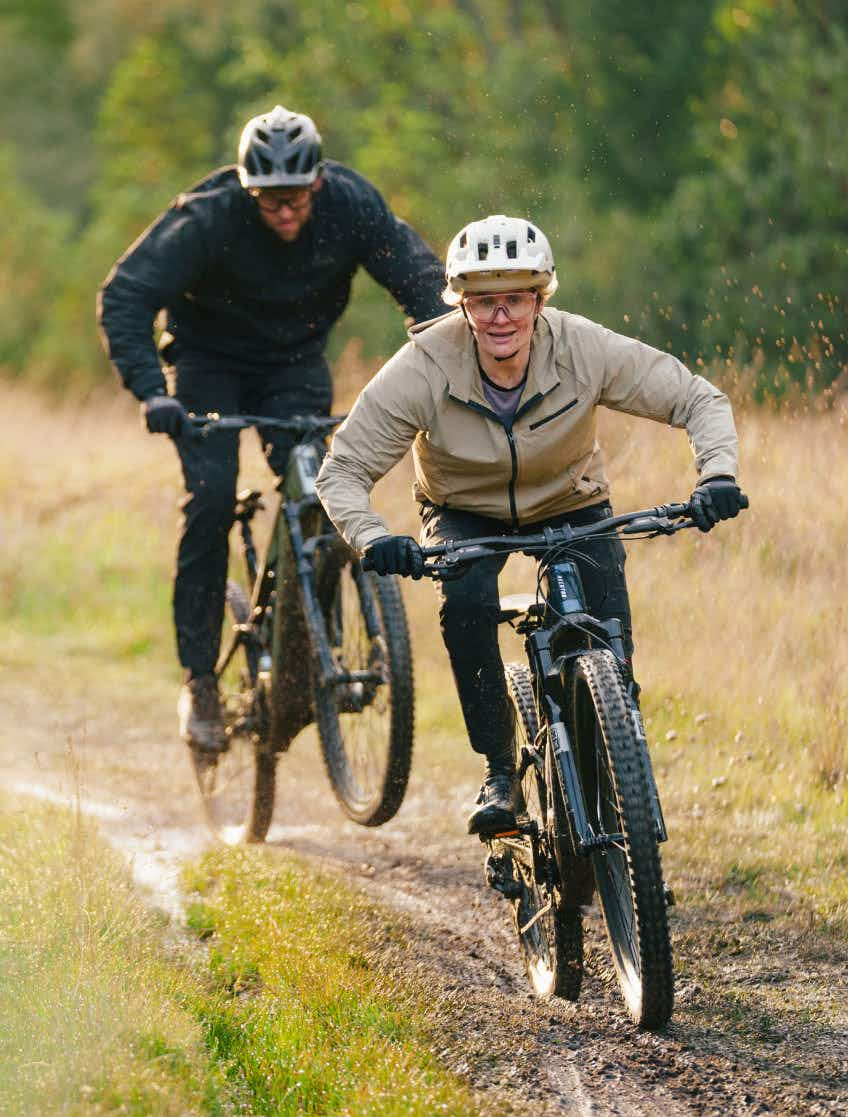 Two people in helmets and jackets ride Aventon Current EXP electric mountain bikes by Aventon on a muddy outdoor trail, one following closely behind the other, with grass and trees in the background.