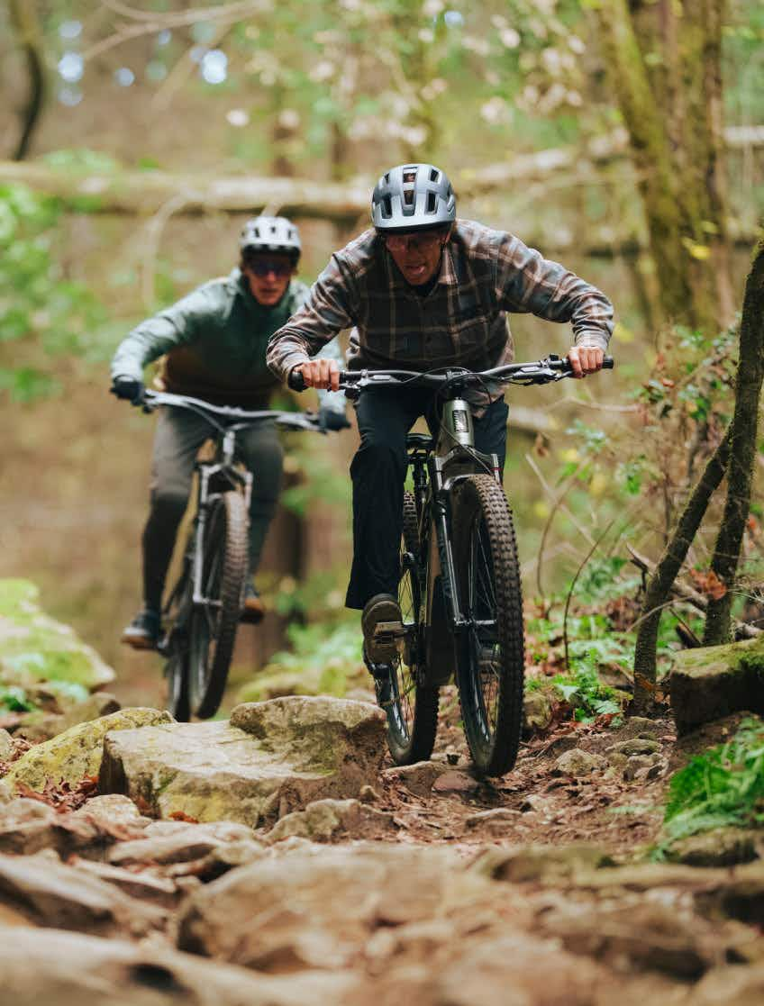 Two people wearing helmets ride Aventon Current EXP carbon eMTBs with mid-drive motors over a rocky forest trail, surrounded by trees and greenery.