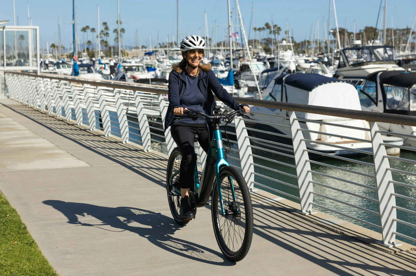 A rider wearing a helmet cruises along a waterfront path near boats and a marina on an Aventon Pace 5 REC Step-Through electric bike by Aventon.