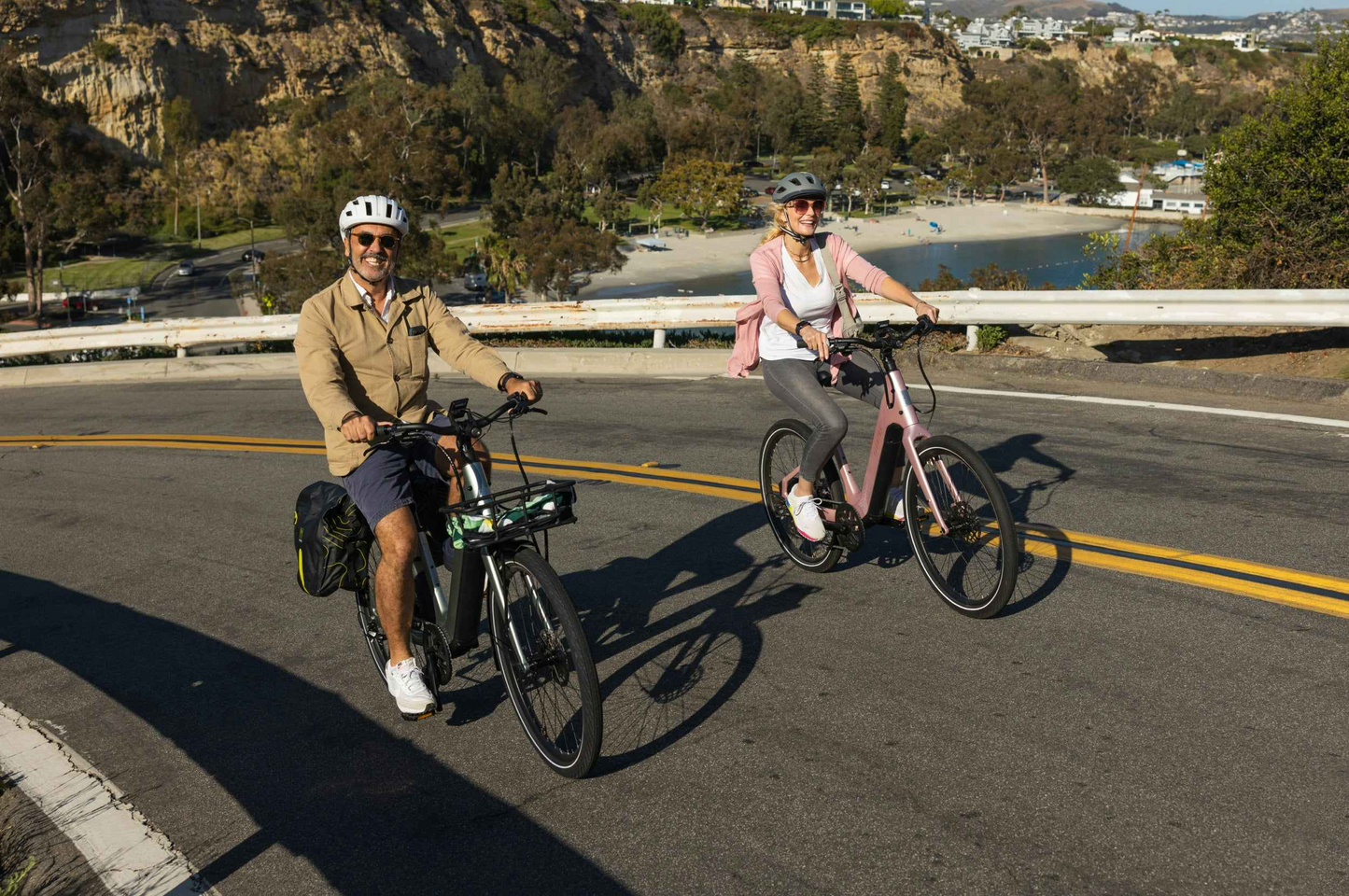 Two adults ride Aventon Pace 5 REC Step-Through e-bikes along a scenic coastal road, enjoying the comfort cruiser experience from Aventon with cliffs and trees in the background.