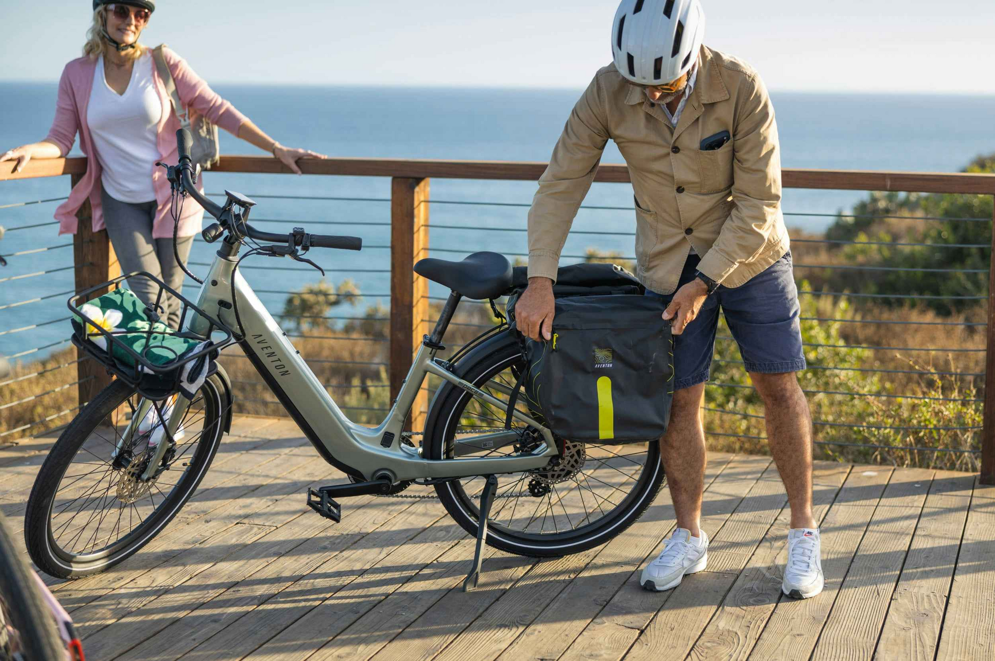 A man in a helmet adjusts a pannier bag on his Aventon Pace 5 REC Step-Through e-bike by Aventon, while a woman stands nearby on a wooden deck with an ocean view.
