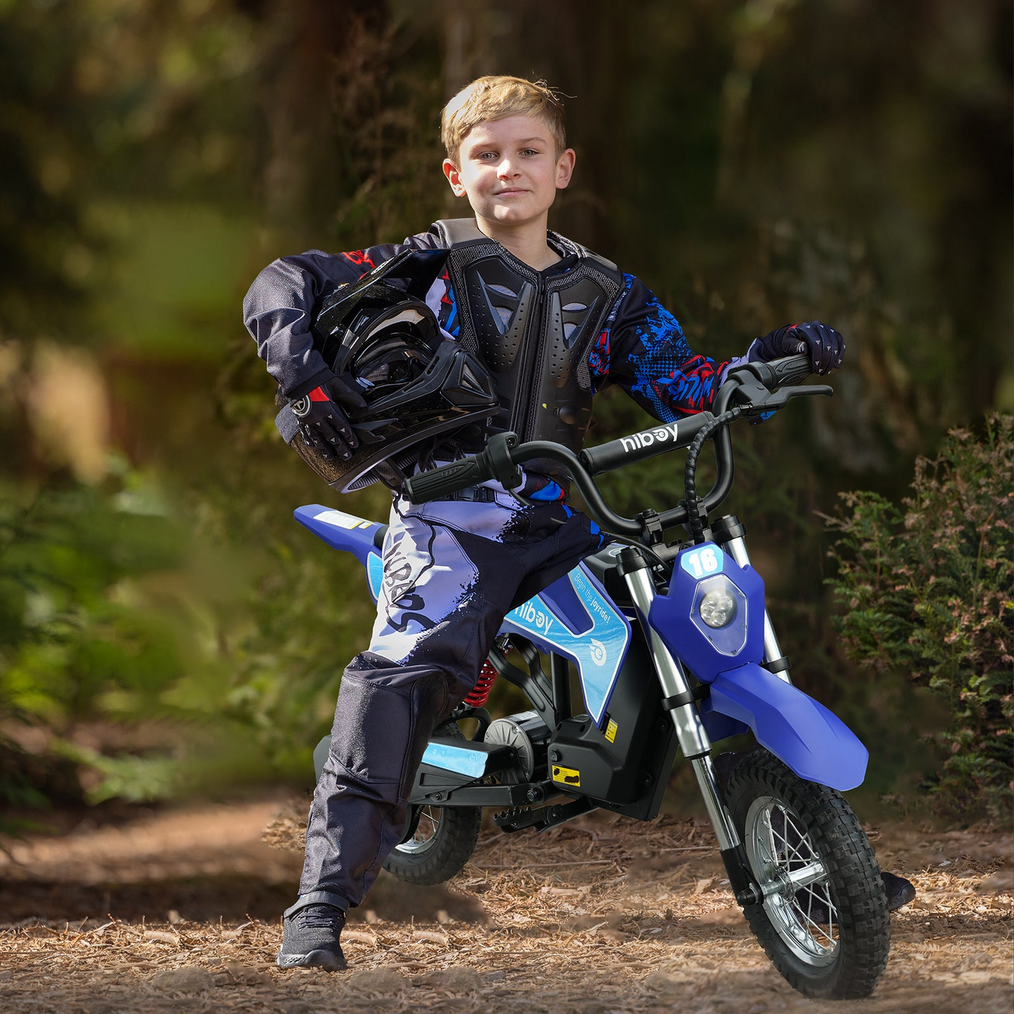 A boy in motocross gear stands beside a Hiboy DK1 Electric Dirt Bike by Hiboy, holding a helmet under his arm in an outdoor wooded area.