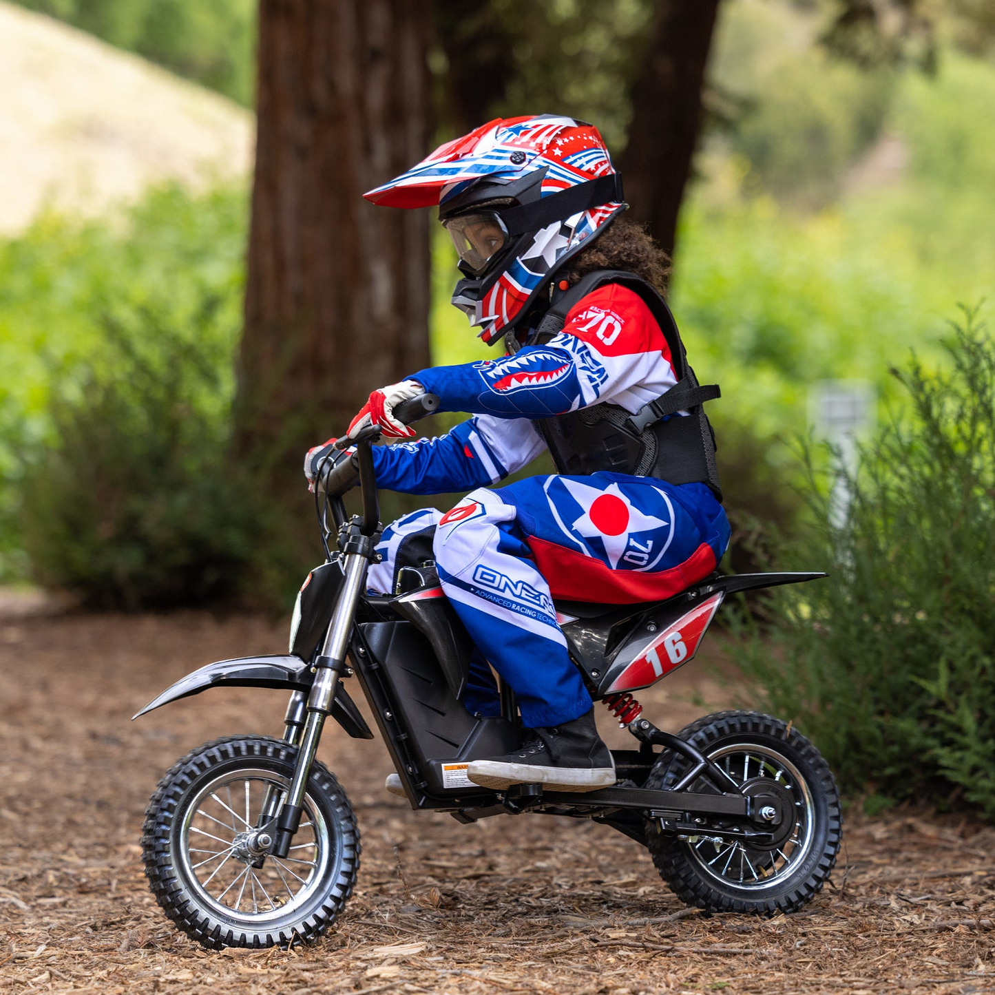 A child in a helmet and protective gear rides the Hiboy DK1 Electric Dirt Bike by Hiboy outdoors, surrounded by trees and greenery.
