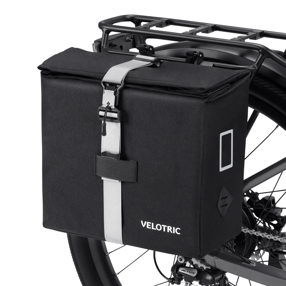 A Velotric Rear Rack Pannier Bag in black with a white strap is mounted on the bike’s rear rack. This Velotric accessory features a buckle closure, visible side patch, and waterproof protection for your gear.