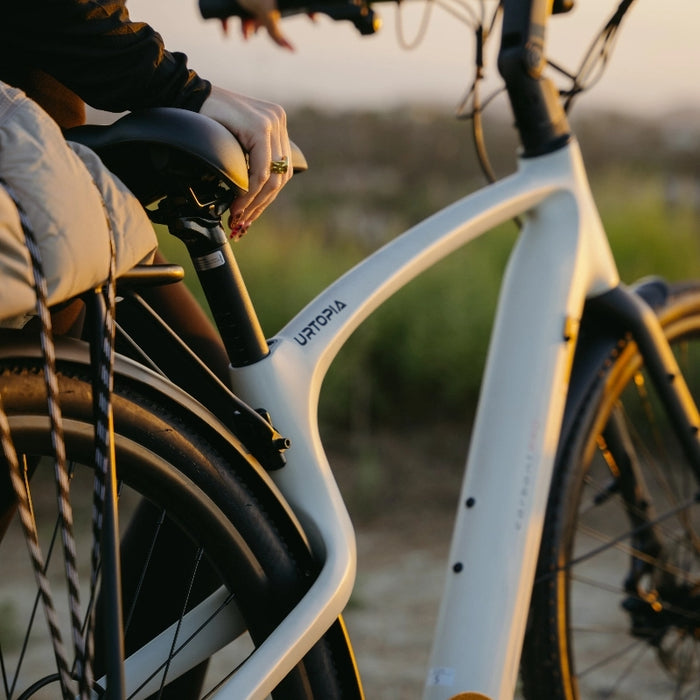 A person rests their hand on the seat of a Urtopia - Carbon Classic by Urtopia, a lightweight electric bike, parked outdoors at sunset with greenery in the background.