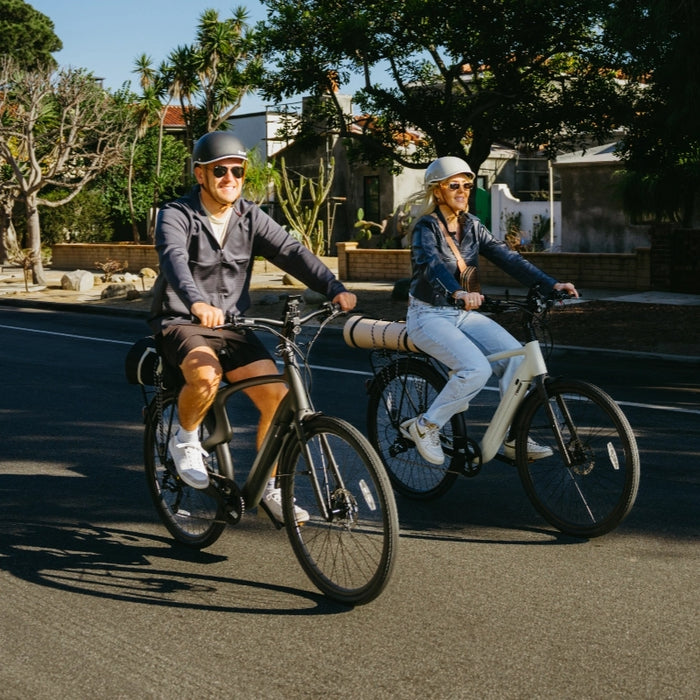 Two people wearing helmets ride Urtopia Carbon Classic electric bikes by Urtopia on a sunny, tree-lined street with houses.