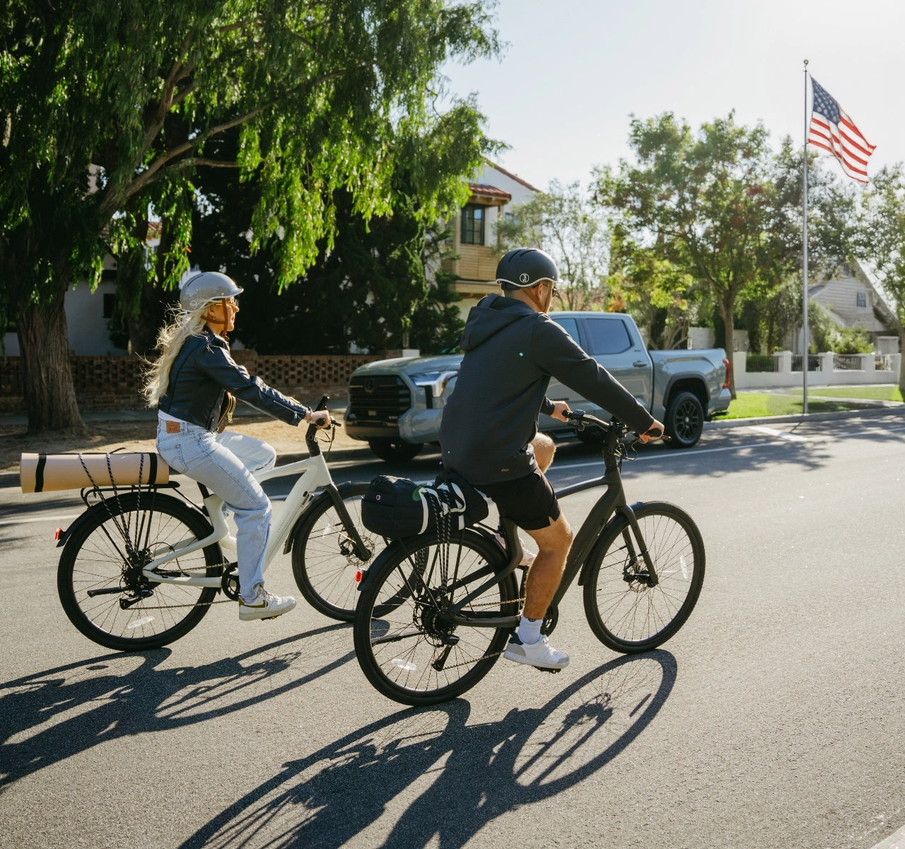 Two people wearing helmets ride the Urtopia - Carbon Classic e-bike by Urtopia on a sunny suburban street lined with houses, trees, parked cars, and an American flag in the background.