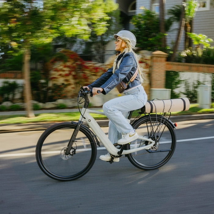 A woman wearing a helmet and sunglasses rides the Urtopia Carbon Classic electric bike by Urtopia, with a rolled yoga mat on the rear rack, along a sunny street.