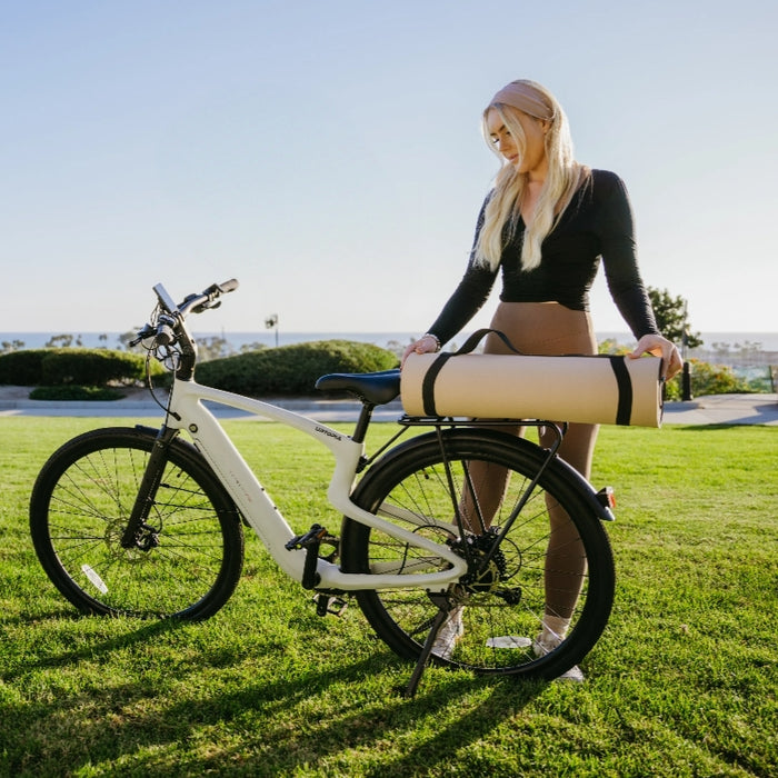 A woman stands on grass beside the Urtopia - Carbon Classic electric bike by Urtopia, holding a rolled-up yoga mat, with trees and the ocean in the background.