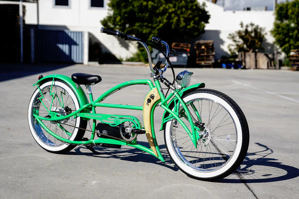 A Tracer Signature Pro 26'' 800W Chopper Cruiser E-Bike, featuring a sleek green design with a long frame and wide handlebars, is parked on a concrete surface. Trees and buildings can be seen in the background.