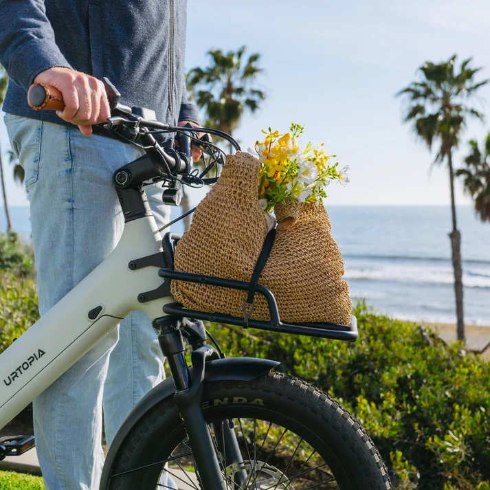 A person stands beside a white Urtopia e-bike with the Urtopia Front Rack - Joy Carbon, filled with baskets of flowers, near the ocean with palm trees in the background.