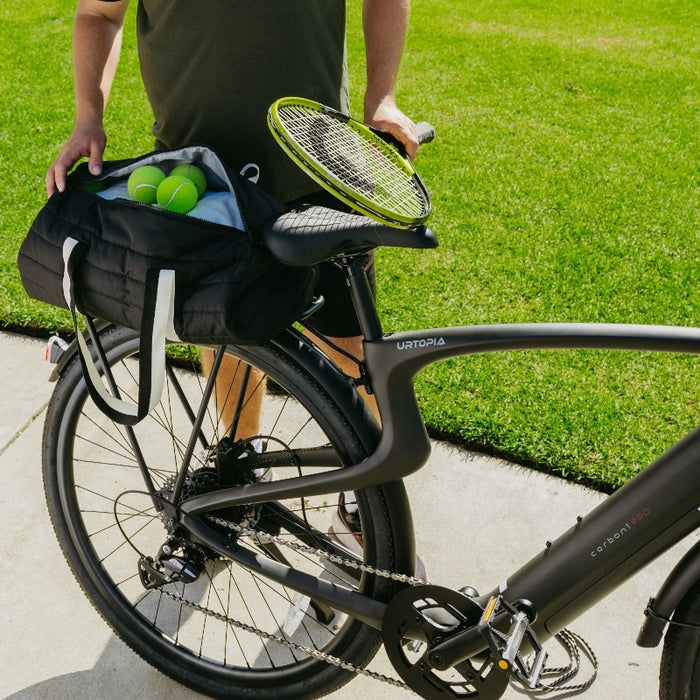 A person stands beside their Urtopia Carbon Classic e-bike, holding the handlebar, while a sports bag with tennis balls and a racket sits on the seat on a sunny day.