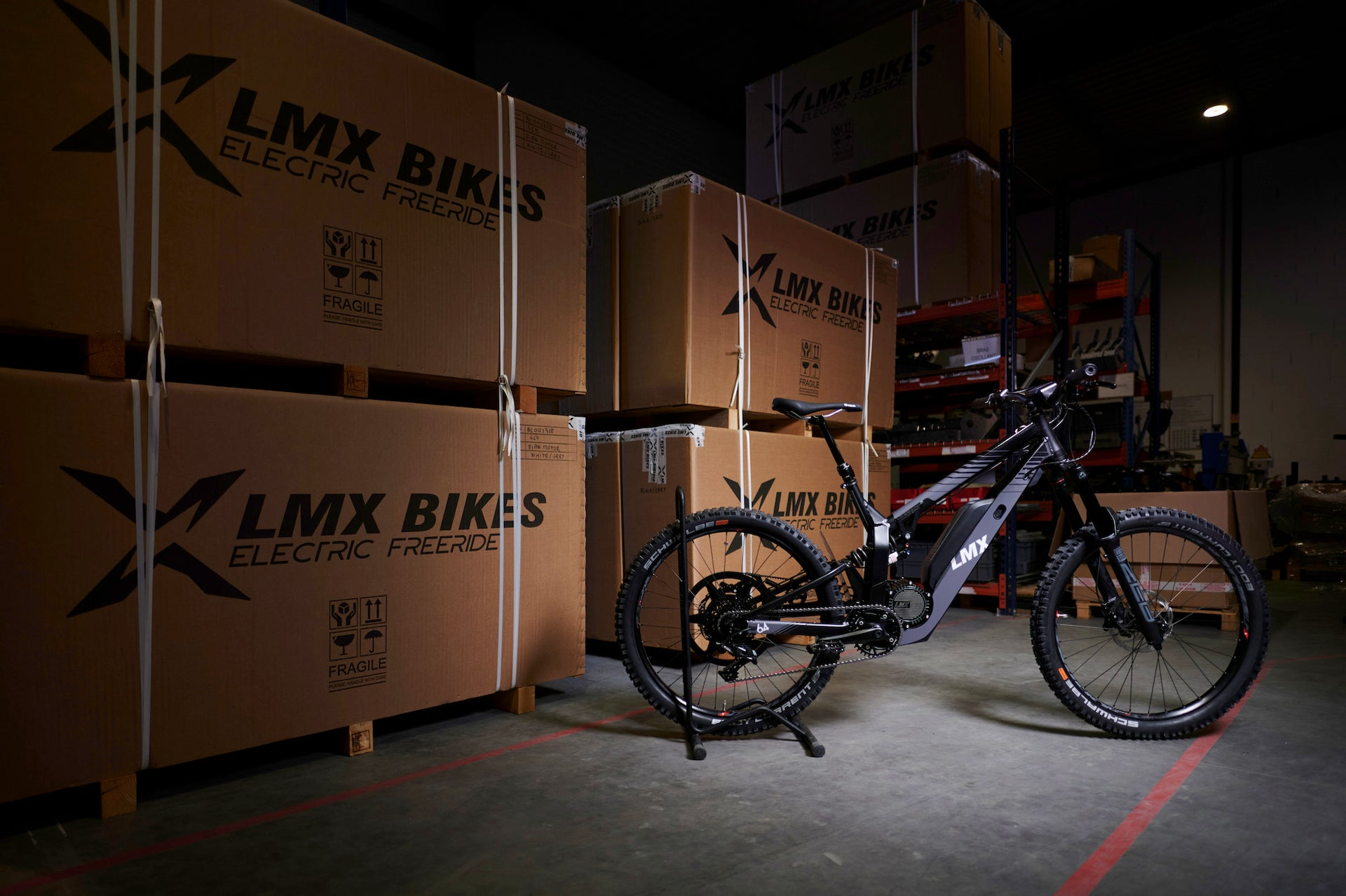 A black electric mountain bike with a brushless mid-mounted motor stands in a dimly lit warehouse, positioned in front of large cardboard boxes labeled "LMX - 64 Electric Freeride.