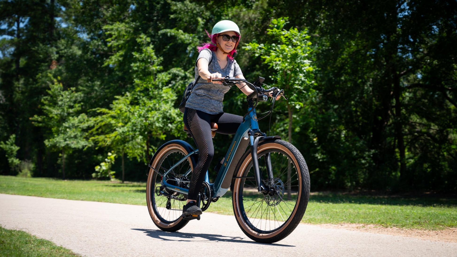 A person wearing a helmet and sunglasses rides a blue AIMA - Santa Monica eBike with a powerful 750W Bafang Motor on a paved path through a green, wooded area.