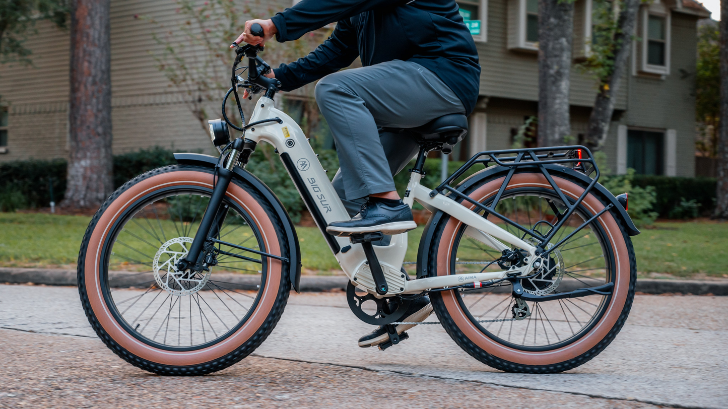 A person rides a white AIMA - Big Sur eBike on a residential street, with houses and trees in the background, effortlessly propelled by its powerful motor.