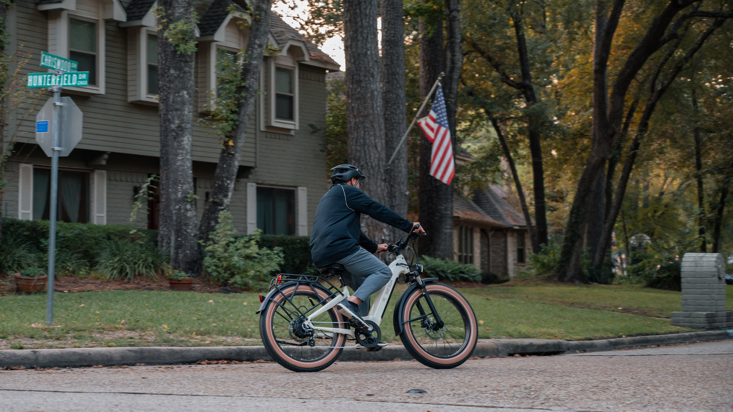 A person rides an AIMA - Big Sur eBike down a residential street lined with trees and houses, with an American flag visible in the background.